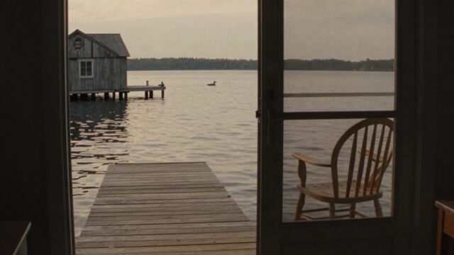 A calm late-afternoon view of a wooden dock on a still northern Minnesota lake, gold light on the water, a weathered boathouse at the shore, a loon's silhouette far out, a single o