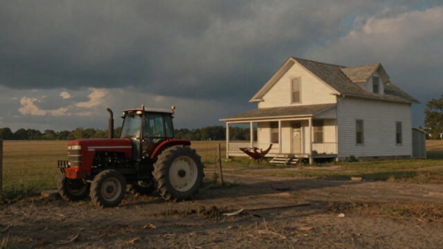 An early evening Missouri farm at the end of August: a dusty north field with a red tractor parked near a barbed-wire fence, a weathered white farmhouse with a wide porch, warm low