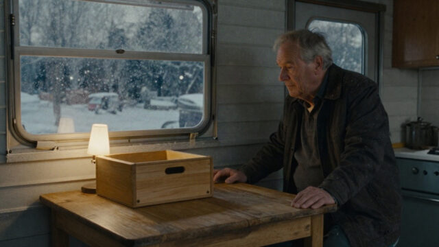 A small kitchen inside a snowy trailer park at dusk on Christmas Eve, warm yellow lamplight spilling across a worn wooden table, a cedar box resting on the table, and snowflakes dr