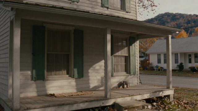 A small wooden front porch of an older two-story house on a quiet mountain street in late autumn, with green shutters, a braided rug visible through a window, and soft afternoon li