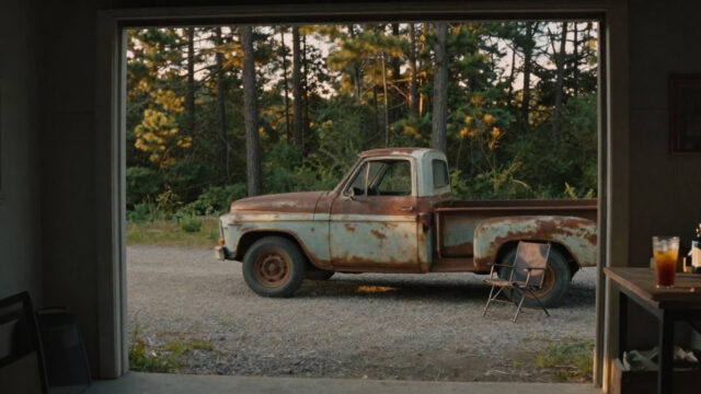 A small country garage at late afternoon in July, the open door framing a gravel drive and tall pine trees. Warm golden light falls across an old rusted pickup truck with its bench